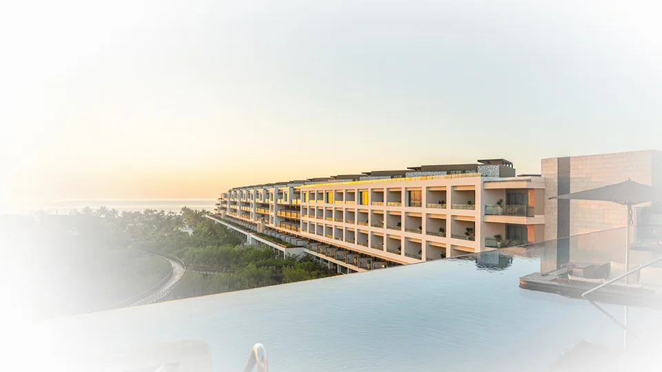 Rooftop infinity pool edge looking along a long, low cream-coloured resort building with balconies at sunrise; heavy white vignette edited onto the edges. No art collection visible.