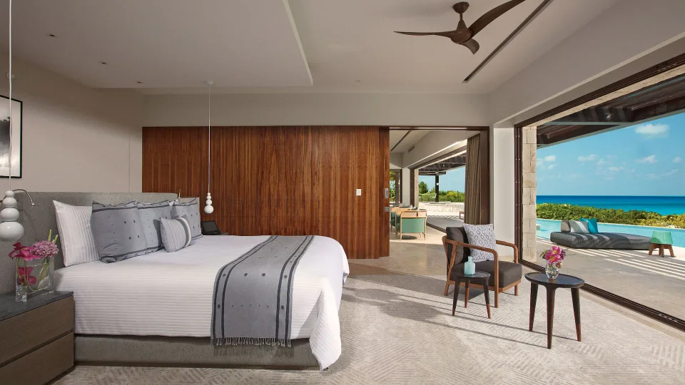 File with no extension (a 1920x1080 WebP): master bedroom with a king bed, grey linens and a walnut-panelled wall, opening onto a terrace with a large infinity pool, daybed and ocean view; it shows a villa-style suite, not a swim-out