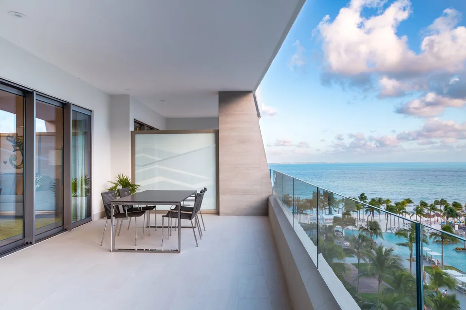 Large covered upper-floor terrace with a dining table for four and a glass railing, looking down on the palm-lined pools and beach toward the open sea. It shows an ocean-view suite terrace on a tower, not a cliff