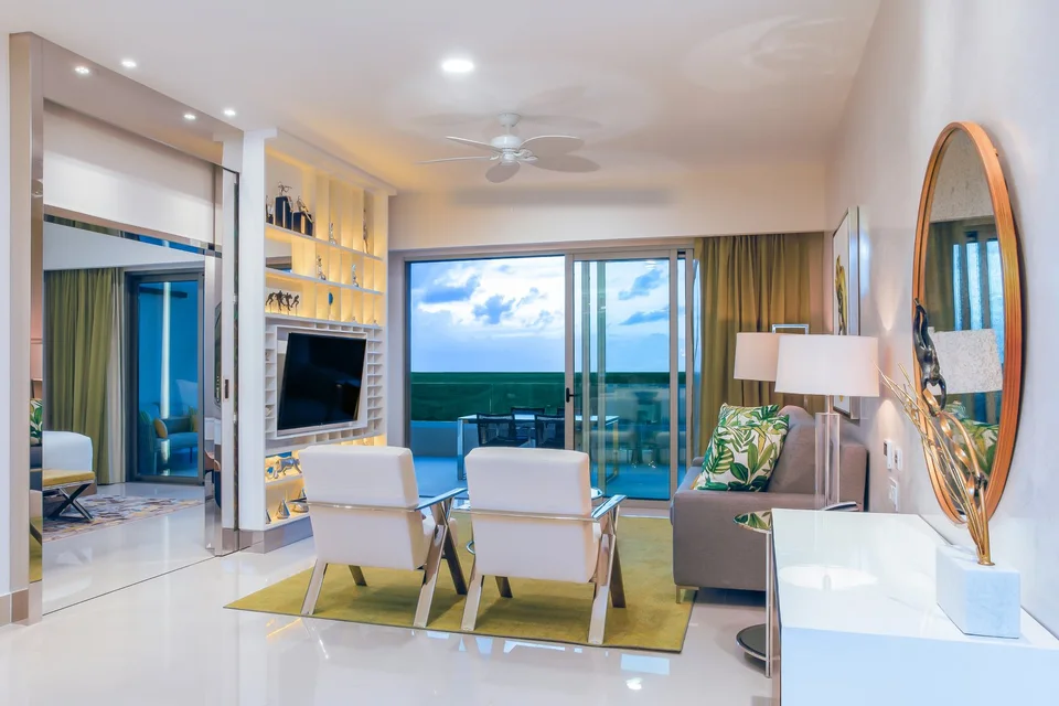 Suite living room in white and chartreuse-green (two white armchairs, grey sofa, TV in a lit shelving wall, round gold mirror), with sliding doors to a terrace with a dining table and a sea view at dusk, and a bedroom visible to the left. It is a one- or two-bedroom suite interior, not a beach cove
