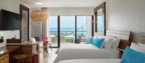 Suite with two double beds, turquoise embroidered cushions, framed folk-art textile panel, woven pendant lamp, and a terrace with hammock facing the sea. Not a 'Muralism Suite' (no such category); category not identifiable from the image.