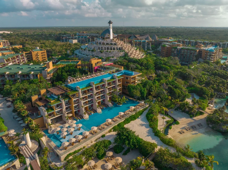 WebP without extension: late-afternoon aerial of orange suite buildings with stone pillars, a long pool with umbrellas, the Xpiral tower behind and inlet beaches to the right. Shows the complex; not a cenote.