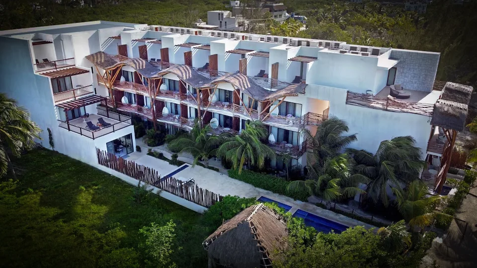 Drone view of the white main building's land side: wooden pergolas and branch-style balcony screens, a narrow pool along the ground floor, palms, and a thatched palapa in the foreground. The beach is not in frame.
