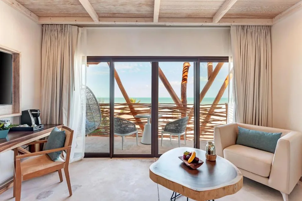Suite sitting area with a live-edge table, an armchair and a desk with a coffee maker, and sliding doors to a balcony with a hanging egg chair, a branch-style railing and a turquoise sea view