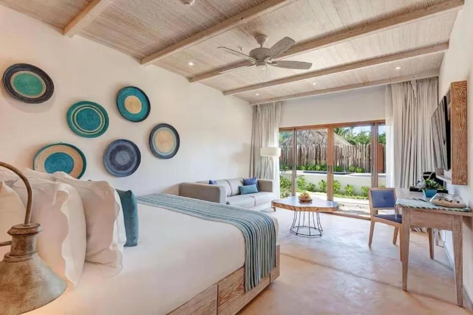 Ground-level suite with a king bed, woven wall plates in blue tones, a sofa and a desk, with sliding doors to a terrace by a pool, a palapa and a wooden fence. Garden or pool side, not the ocean.