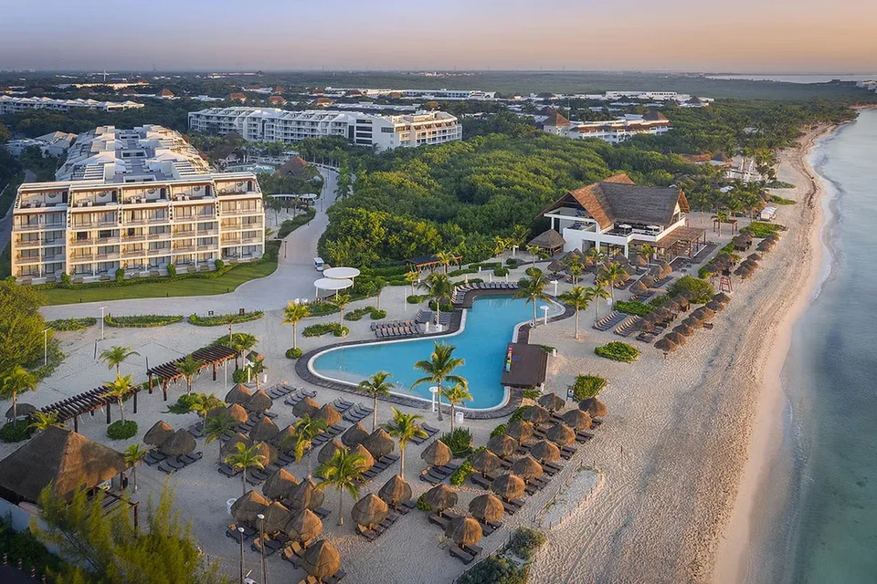 Official aerial at sunrise or sunset: beachfront pool, rows of palapa sunshades on the sand, a thatched beach restaurant and the resort's white accommodation blocks (official 'Aerial view of the hotel'). The .heif file is the same image.