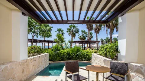 Pergola-covered terrace with stone walls, a small private plunge pool, a bistro table and two chairs, with gardens and beach palapas behind; it looks like a swim-out terrace, but the category is unconfirmed