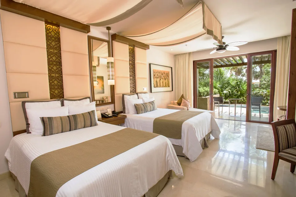 Junior suite with two double beds under fabric canopy drapes and ornate headboard panels, opening onto a garden terrace with a small pool. This is the older decor, which predates the 2025 redesign; nothing shows Preferred Club