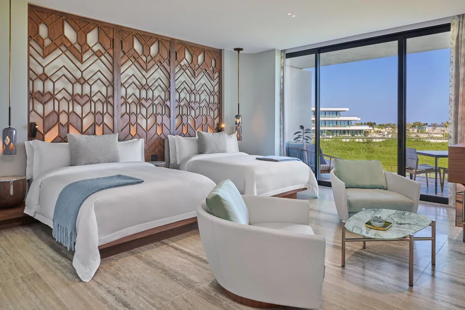 Guest room with two queen beds, carved walnut lattice headboards, two armchairs and a sliding glass door to a terrace overlooking the mangrove, with a resort building behind. No sea view in frame.