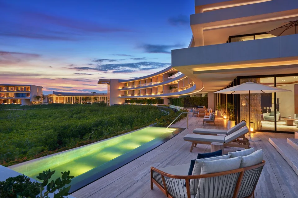 Terrace at dusk with a long lit private pool along the mangrove edge, loungers, an umbrella and a glass-walled living room, with the curved buildings lit in the background. No ocean visible.