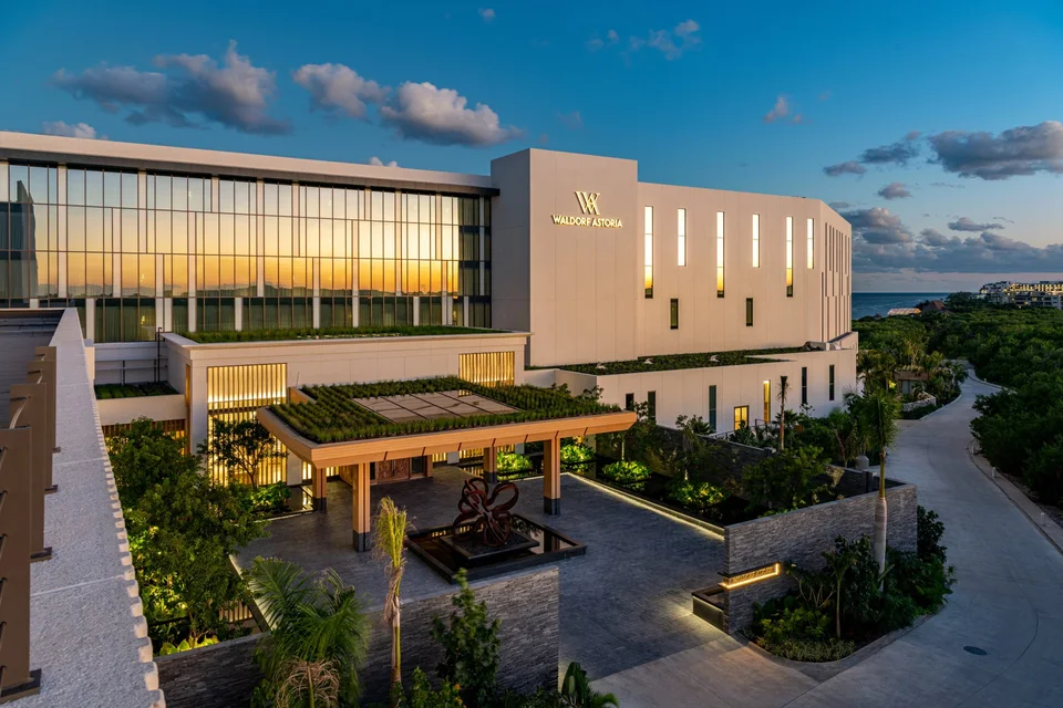 Exterior at dusk: the arrival court and porte-cochère with a green roof, a red flower sculpture in a reflecting pool, and the lit 'Waldorf Astoria' sign on a white building, with the sea in the distance. Not a lobby interior. Identical to media/originals/hotels/waldorf-astoria-cancun/hero.jpg.