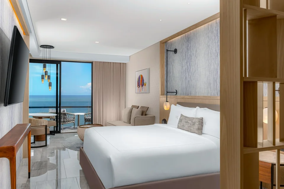 King bedroom with a WA-monogrammed pillow, a sofa and dining table, and a sliding door to an ocean-view balcony with chairs