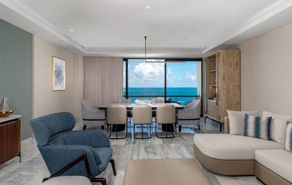 Suite living and dining room with a sectional sofa, a blue armchair, a table for eight and floor-to-ceiling windows onto the sea. It looks like a suite living room, not a junior suite terrace. The eight-seat table fits Hilton's description of the Deluxe Ocean Front Suite, but this is not confirmed.