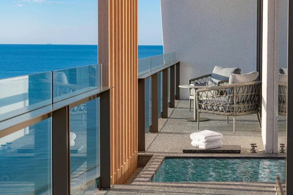 Upper-floor balcony with a glass railing, a small in-deck pool or soaking tub, rope armchairs and slatted wood, looking over the open Caribbean. It shows the ocean, not a lagoon.